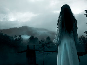 A long dark-haired woman in a long white dress looks out over a foggy, desolate landscape tinted blue.